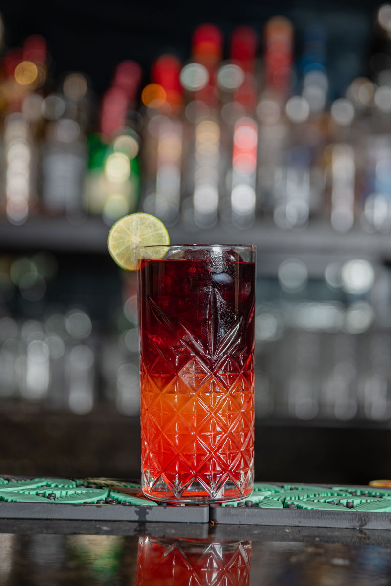 Coctel de tono rojizo con rodaja de lima frente a las botellas de la barra